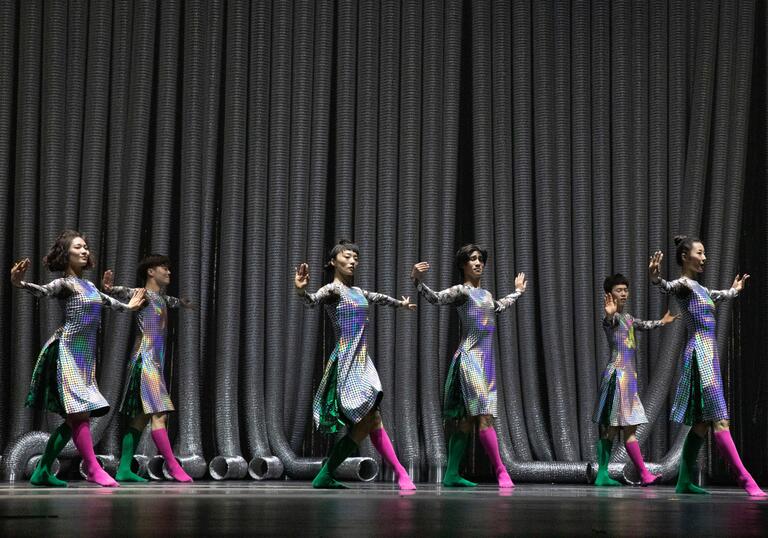 Six performers stnad in pose wearing silver dresses and lock pink socks.
