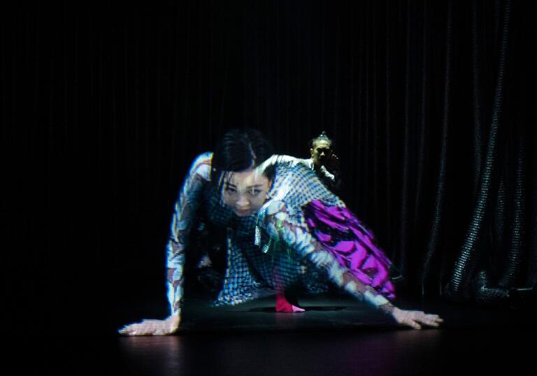 Two performers seem to be merged together while dancing on a stage. One looks like a hologram.