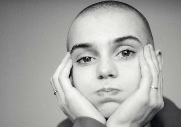 Black and white image of artist Sinead O'conner staring into the camera lens with hands either side of cheek.