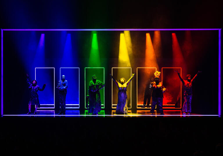 Six performers stand in front of doorways holding poses. Beams of colourful light illuminate them. 