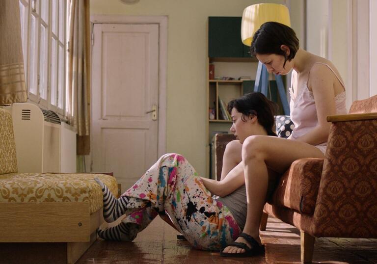 A person sits on the floor of a living room in between the legs of another person sitting behind them on a sofa. A still from A Room of My Own.