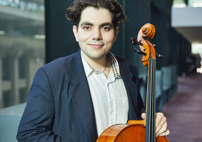 Alex Ciulin holding cello in foyer of Milton Court, with windows behind him looking out to Barbican Estate