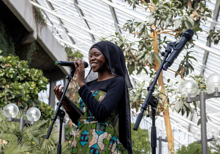 Person in black headscarf and green dress stands with microphone in the Conservatory