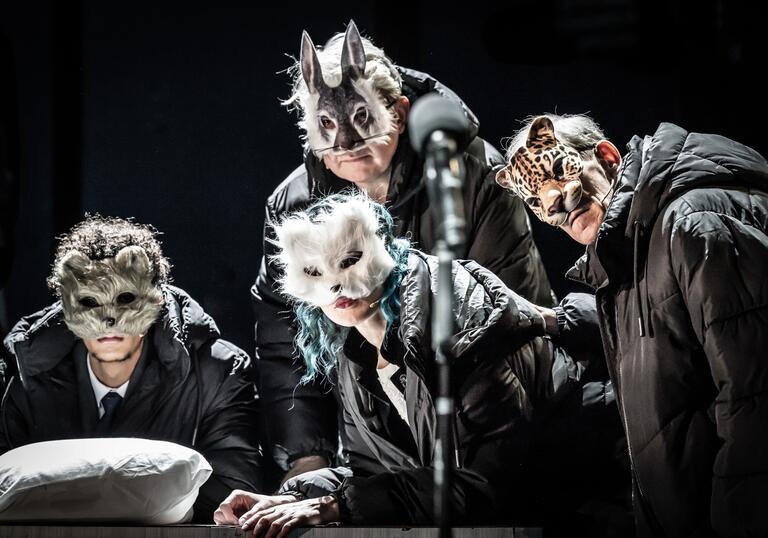Four people wearing black coats and animal face masks gather together as if listening to or watching someone.