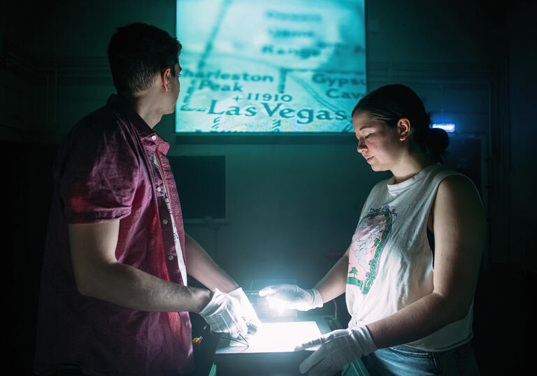 Two people stand in front of a projector wearing white gloves and handling a piece of equipment. An image of a map of Las Vegas is projected behind them.