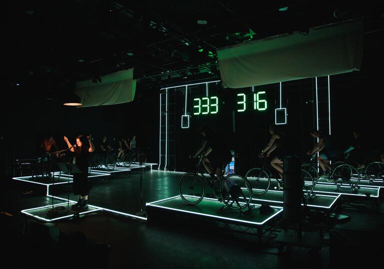 Inside the rehearsal room for A Play for the Living in a Time of Extinction: 10 people power bikes on an electric green set.