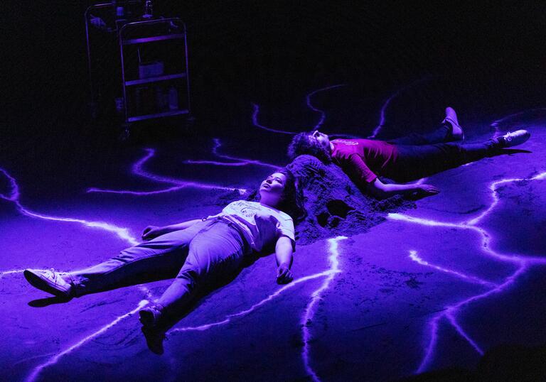 Two people lie on the floor in a dark room. They lie against a mound, which has bright lines coming out of it. They are lit with a purple light.