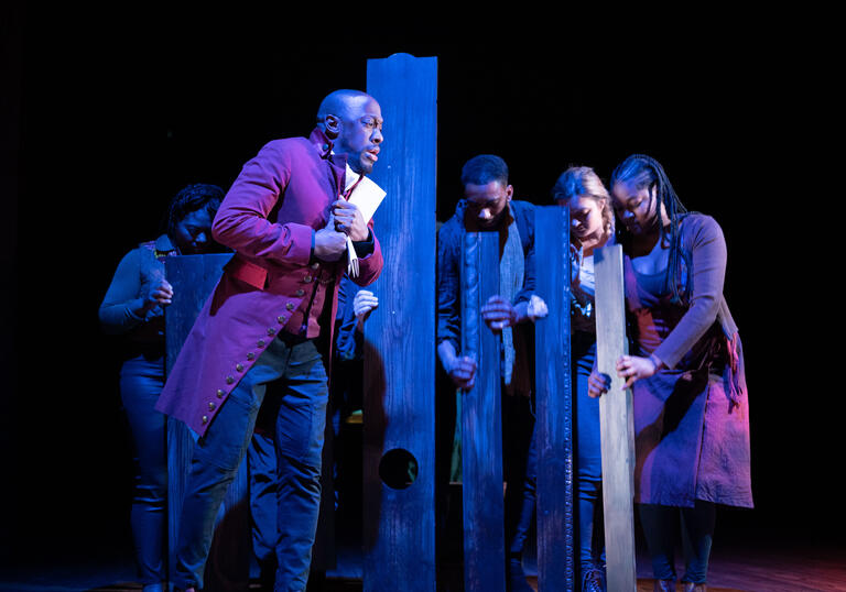 Actor Giles Terera leans against a wooden post while other performers stand with their heads down. 