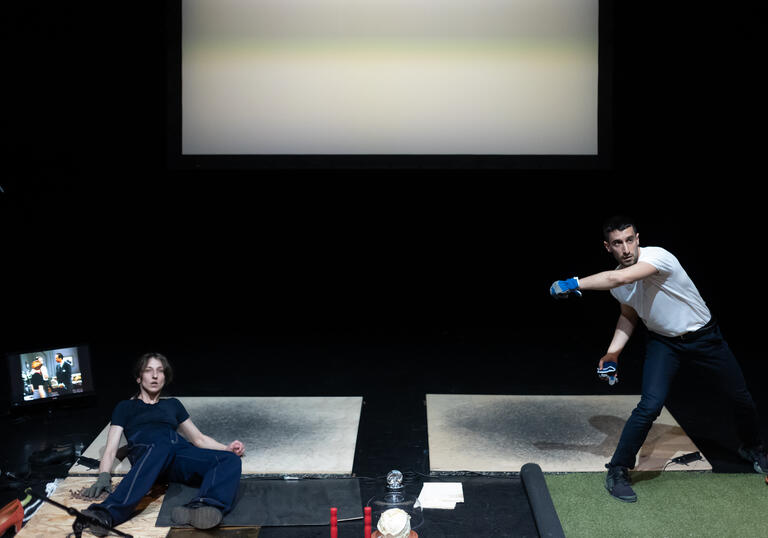 Two performers on stage, one wears gloves and moves around on a piece of fake grass, while the other lies on the floor watching.