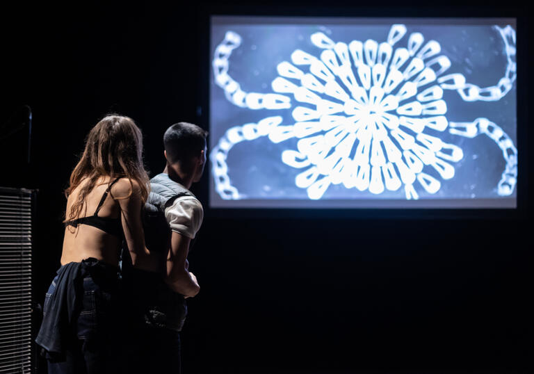 Two performers stand before a screen with a projection of surreal chains forming a circle.