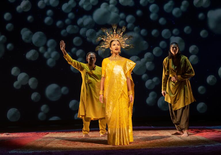 A performer wearing a gold crown sings, while two other performers stand behind.