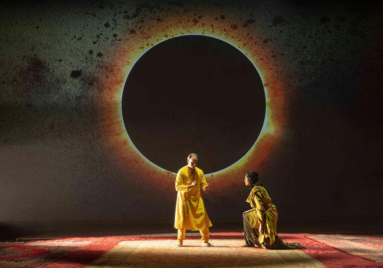 Two performers on stage, one kneels before the other. Behind them, an eclipse is projected,