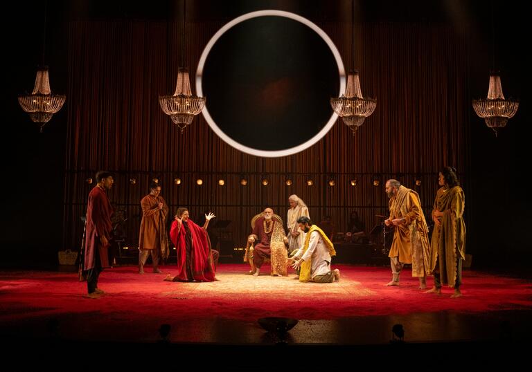 An ensemble of performers gather on stage in a decadent room, a large circle light hangs above them.