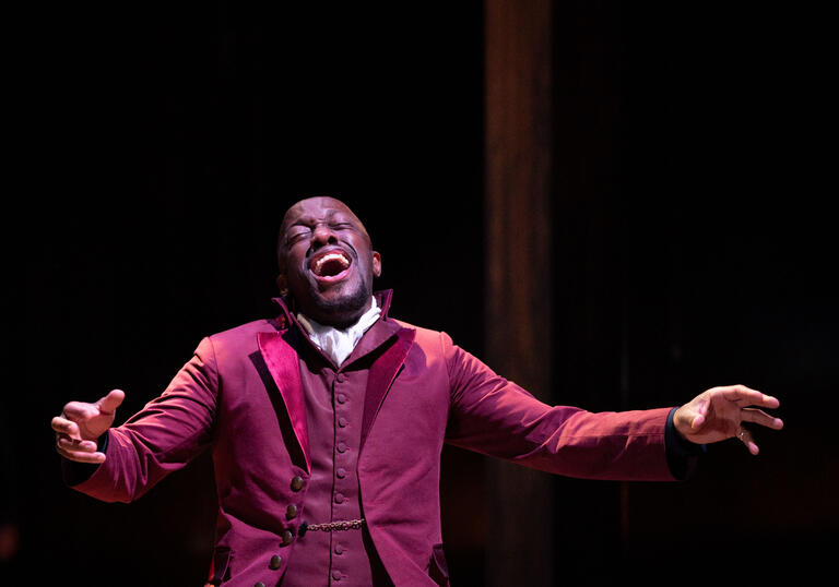 Actor Giles Terera sings profoundly with his arms open while kneeling on stage in a red jacket