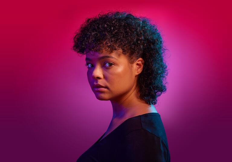 Actor Lydia West stares down the camera while standing against a pink background. The world is on fire in her eye.