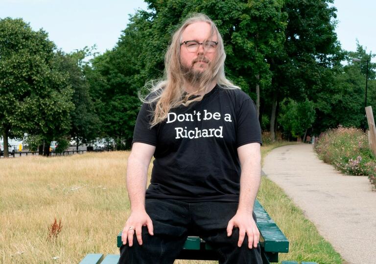 Richard Dawson sitting on a park bench with and ironic t-shirt sloganed with "Don't be a Richard"