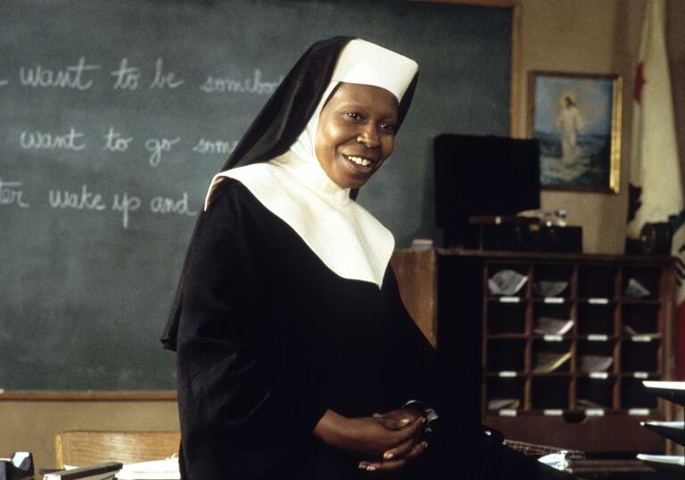 A still from the film Sister Act 2: Whoopi Goldberg is dressed as a nun and sits on a desk in a classroom smiling.