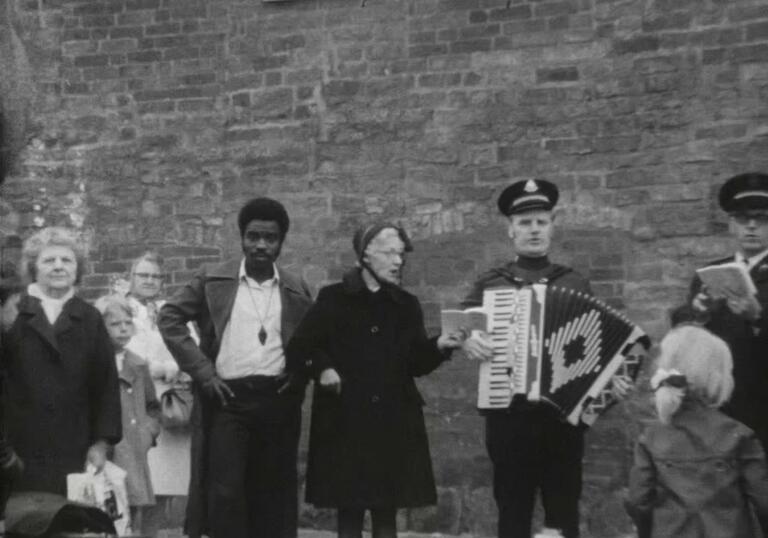 A group of people stand on a stage in the street, reading and playing accordion