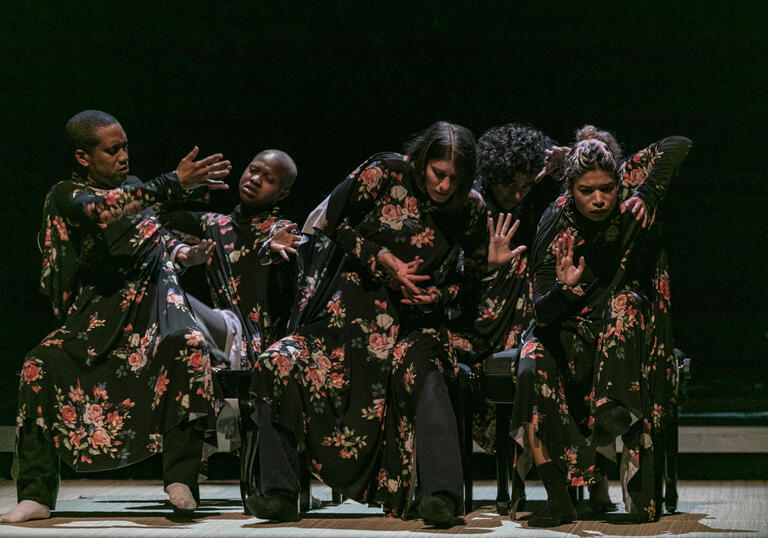 A group of performers wearing long black shawls with red and pink flowers on them move in a group