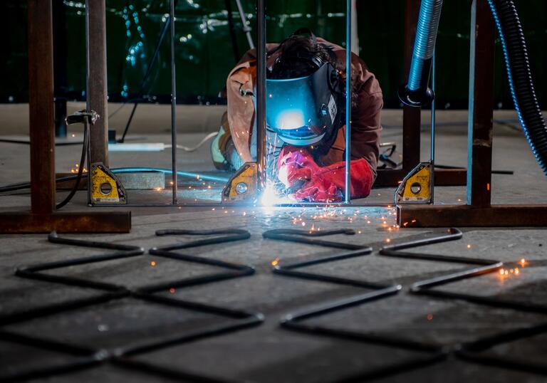 Performance artist Rachel Mars lies on the floor melding a metal gate. She wears protective clothing, including a metal helmet.