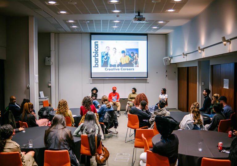 Group of people sitting in a room facing a screen and 4 people people sitting on a panel