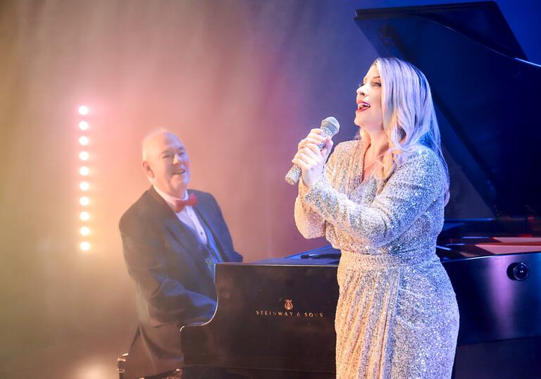 Louise Dearman singing into a microphone, with pianist Jonathan Cohen on piano behind her