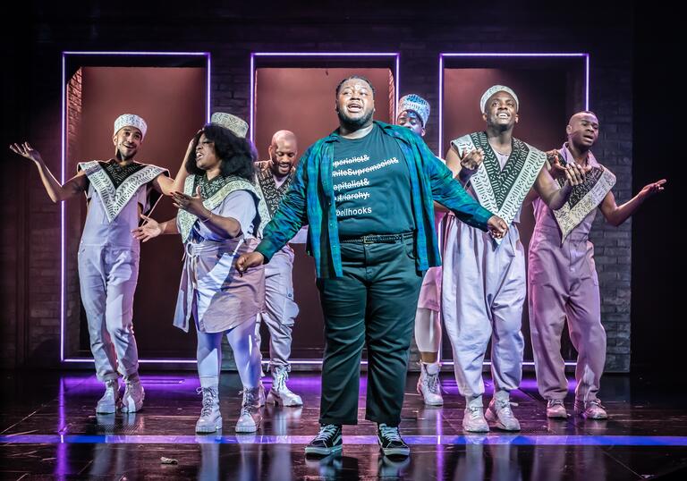 The company of a strange loop pose on stage. A person wearing a green and blue chequered shirt stands front and centre, as six other performers sing and pose behind them.