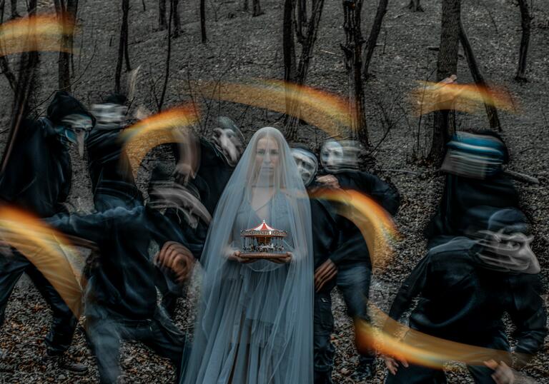 A lady in long kthin light blue veil is staring at the camera. She is holding a small model of a red and white carousel in her hands. Behind her seven masked figure and blurred as they move, they are holding flares. 