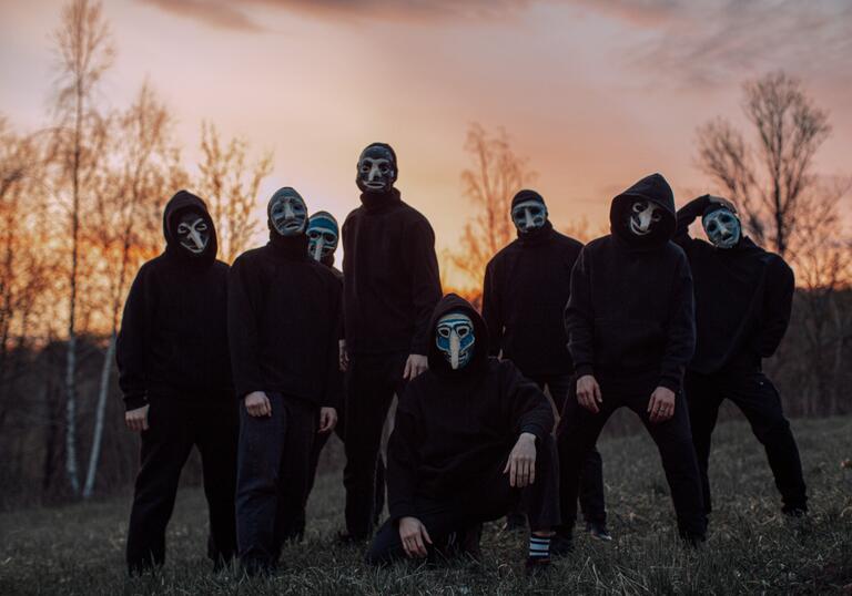 Eight people dressed in black stand in a group in front of a sunset in a field. They are wearing face masks. 