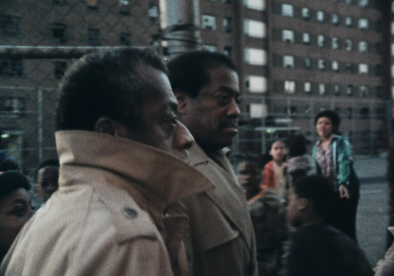 James Baldwin stands on a built up road with his jacket collar around his face