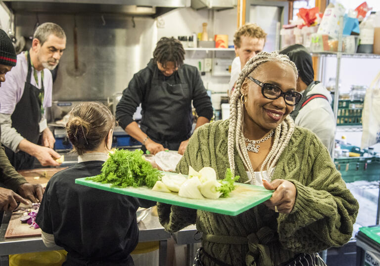 Individual wearing glasses and green cardigan holds tray of ingredients in kitchen