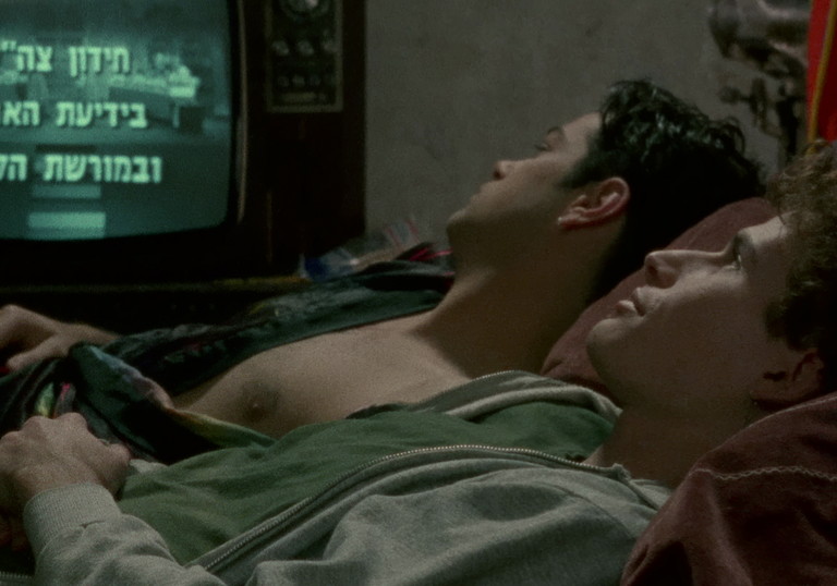 Two men lying in bed together in a dark room look at credits on an old TV screen.