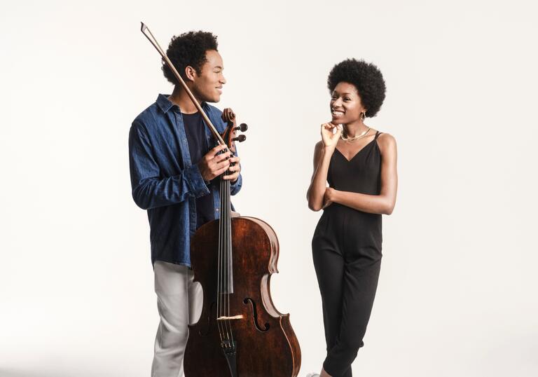 Sheku and Isata Kanneh-Mason smiling at each other. Sheku is holding his cello and bow and they are standing in front of a white background.