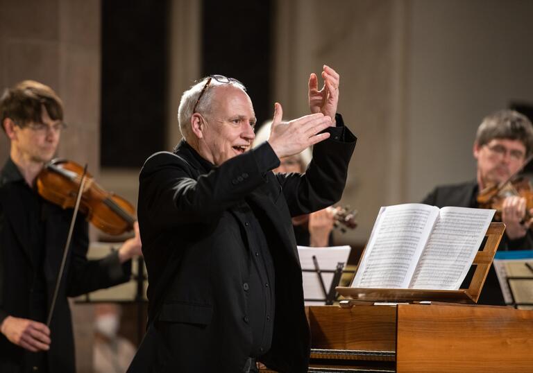 Musical Director John Butt standing by a harpsichord with his arms raised. In the background are two violinists.