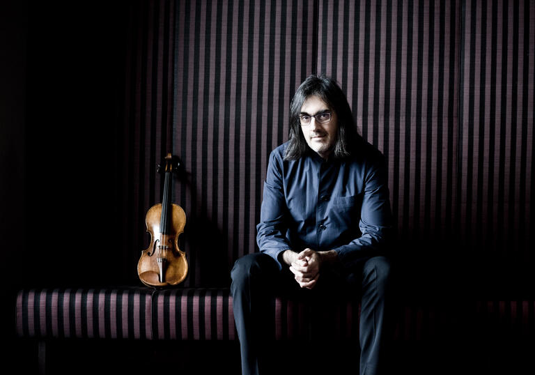 Leonidas Kavakos with his hands clasped sitting on a bench, his violin standing upright next to him