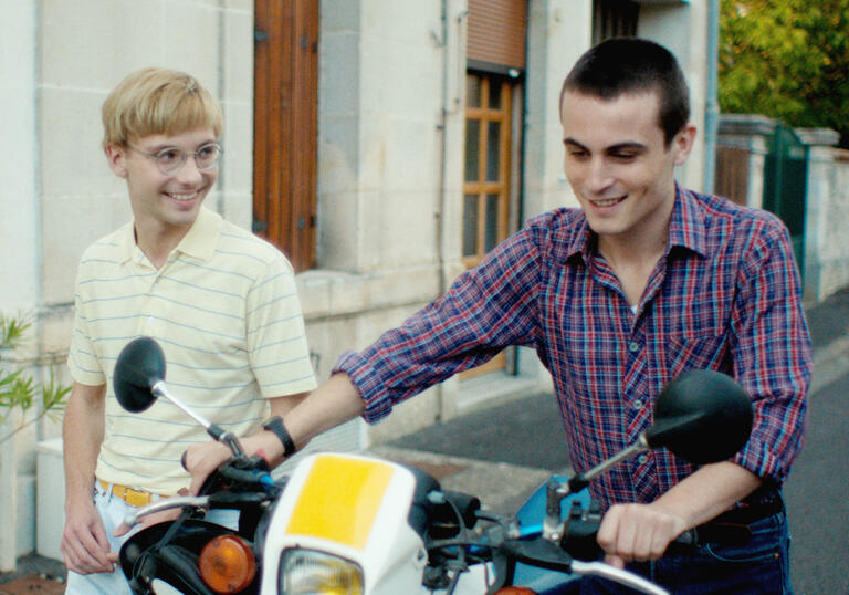 two boys walk down the street together, one pushing a bicycle