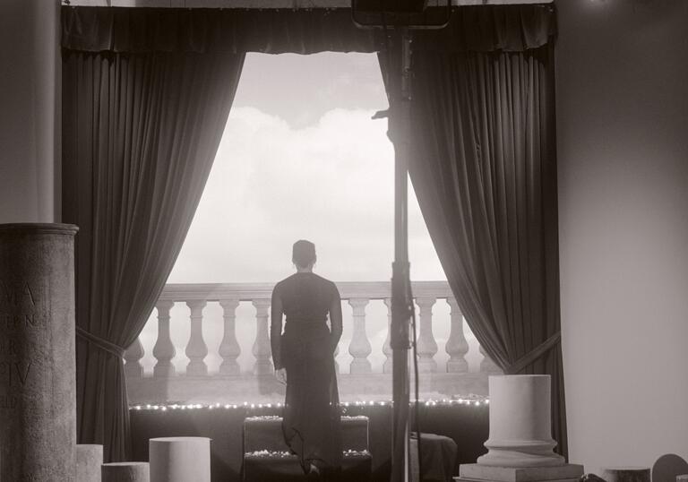 Black and white image of a woman standing with her back to the camera, looking out on to a balcony