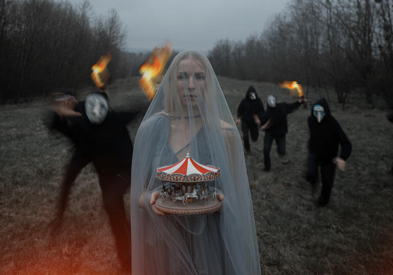 A person wearing a grey veil stands on grass holding a miniature carousel while a group of people dressed in black and wearing haunting facemasks run forwards holding fire.