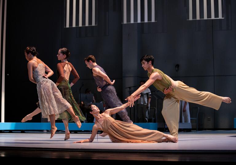 An ensemble of five dancers move and pose elegantly in muted clothing. At the forefront of the stage, two dancers clasp hands. 