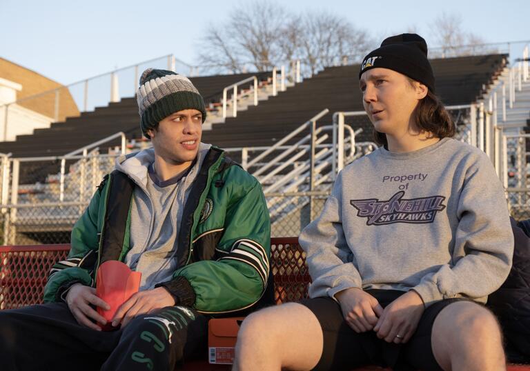 Two young men sit on bleachers looking at each other.