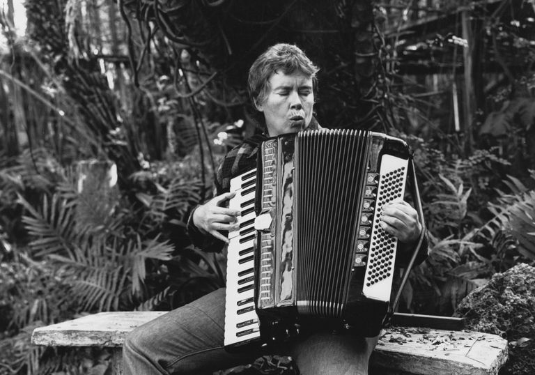 A woman sits on a bench outside and plays an accordion.