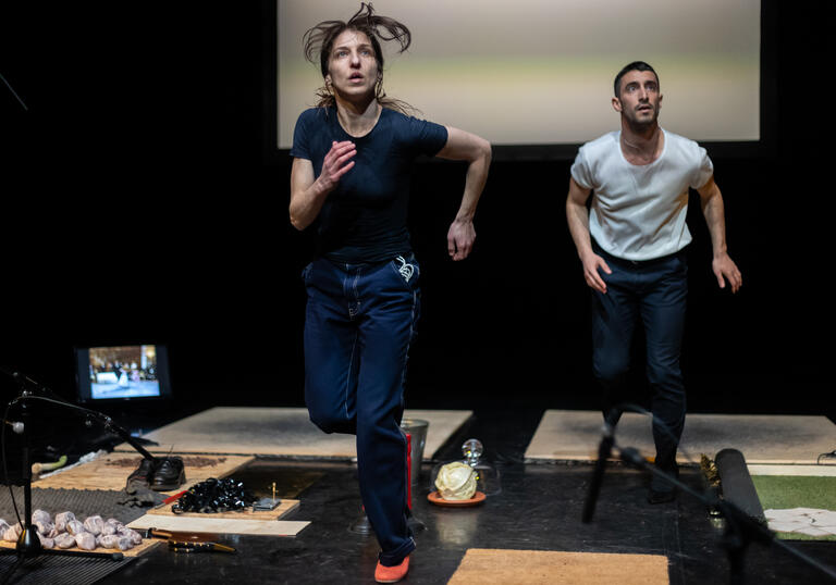 Two dancers run on treadmills... they look out at the audience, watching something, concerned.