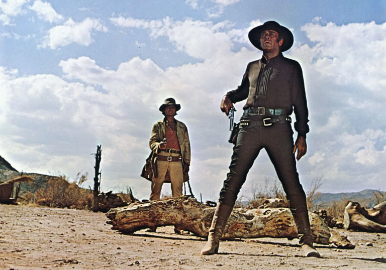 Two cowboys stand in the desert facing the same direction.