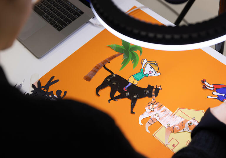 A table with an orange piece of paper has some printed and cut out animated animals which are being arranged, under a bright light.
