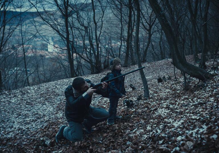 A dad points a gun forward whilst standing next to his young son in a forest at nightfall.