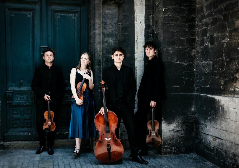 The Sonoro Quartet standing in front of a stone wall and large blue door, holding their instruments