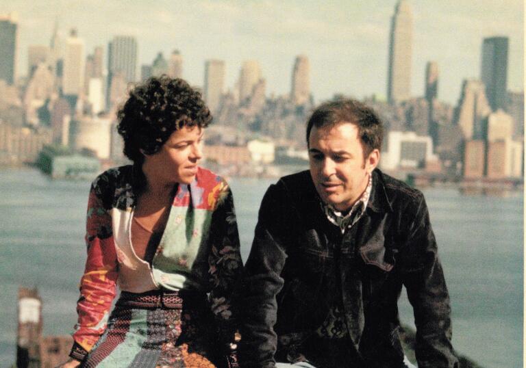Two people sit on a wall in front of a city skyline.