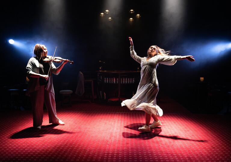 A person in a white dress and trainers dances on a red carpet in a dark, smokey room. To their left, a person dressed all in gray plays the violin.