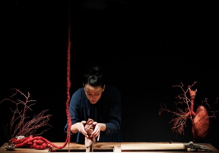 A person wearing blue overalls sculpts a small body out of clay on a table. There is a heart to their left-hand side and a red rope that mimics an intestine.