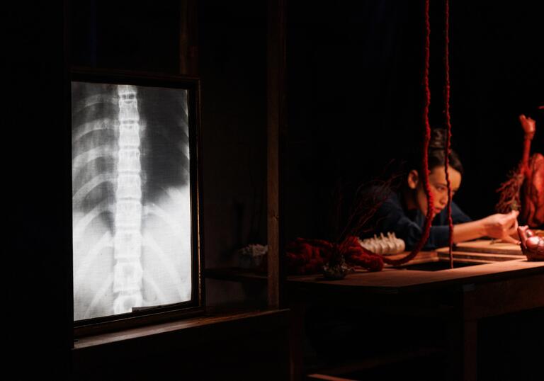 A person wearing blue overalls puts final touches onto a red heart sculpture which hangs above a wooden table. Next to the table there is an x-ray of a set of ribs.